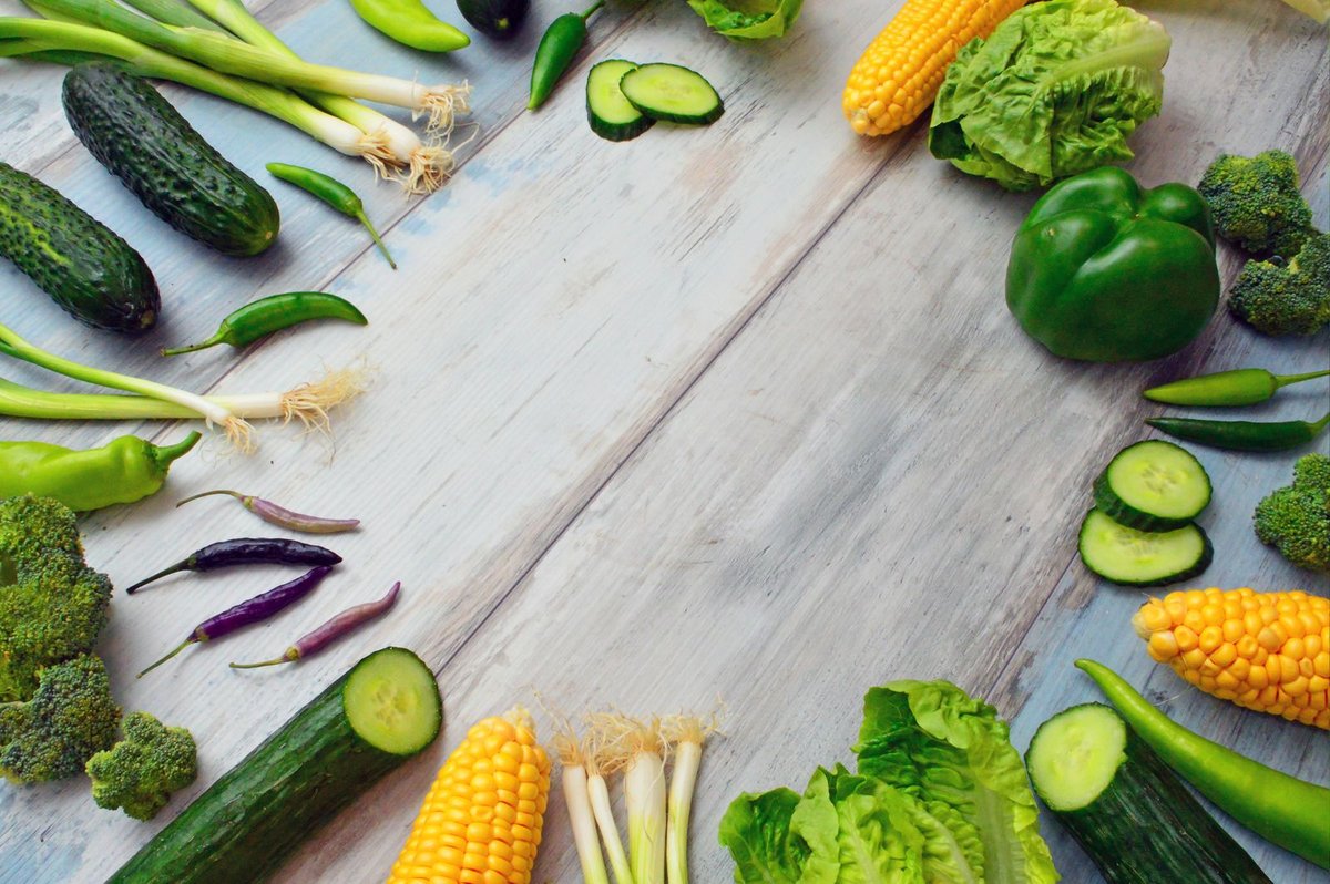 Circle of fresh vegetables including lettuce, corn, cucumber, broccoli, and spring onions on a wooden table